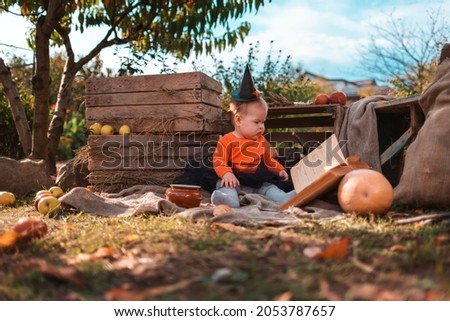 A cute little girl in a witch costume sitting in kitchen-garden and plays with book. Halloween holiday concept.