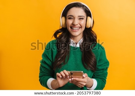 Portrait of a happy smiling casual woman standing over yellow background listening to music with headphones holding mobile phone