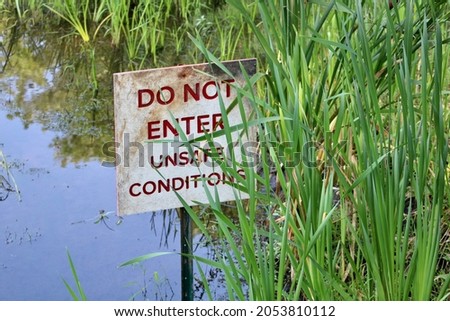 A small, square, metal sign with red, bold, font that reads "Do not enter unsafe conditions". The sign is attached to a metal stake that is sitting in a body of water and hiding behind blades of grass