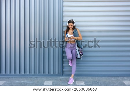 Music and sport. Smiling muscular young woman with headphones, smart watch and bottle of water looks at smartphone on wall background, free space. Athlete runner in sportswear relaxing