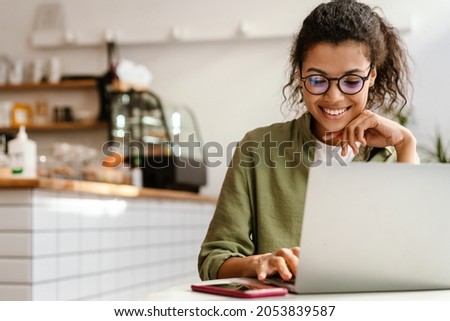 Young black woman in eyeglasses working with laptop while sitting at cafe indoors Royalty-Free Stock Photo #2053839587