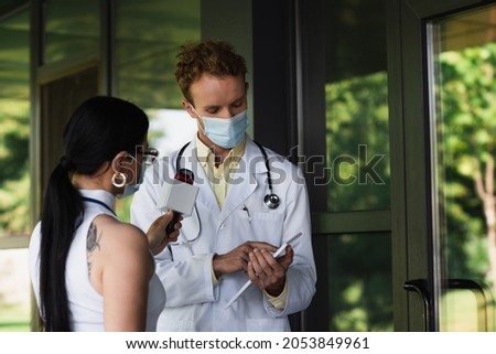 doctor in medical mask pointing at digital tablet near tattooed journalist with microphone Royalty-Free Stock Photo #2053849961