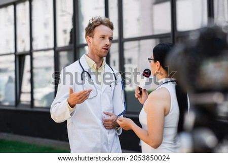 doctor in white coat gesturing while giving interview to asian journalist with microphone Royalty-Free Stock Photo #2053850024