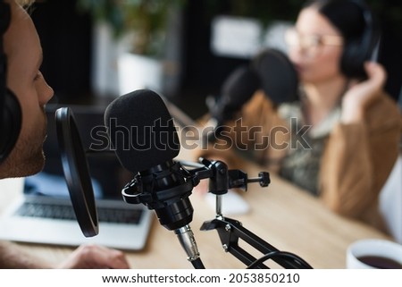 radio host in wireless headphones talking in microphone near blurred asian colleague