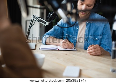 radio host in wireless headphones writing in notebook near blurred colleague