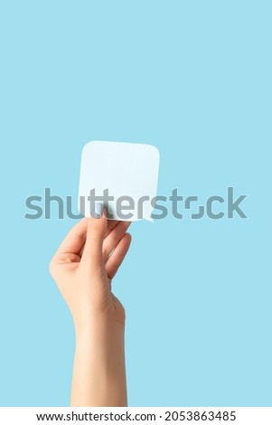 Female hand with blue manicure holding a white blank message card. Speech shape banner in female hand on blue background. Copy space.