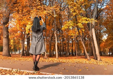 Girl in a coat with long black hair takes pictures on a smartphone of a beautiful autumn park. Autumn trees with golden foliage. Woman in autumn park.
