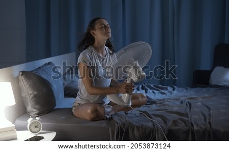 Woman in her bedroom on a hot summer night, she is enjoying fresh air in front of a fan Royalty-Free Stock Photo #2053873124