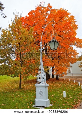 A beautiful street lamp against the background of a maple tree with orange, bright, autumn leaves and a  sky with clouds in the park on the Elagin Island of St. Petersburg.