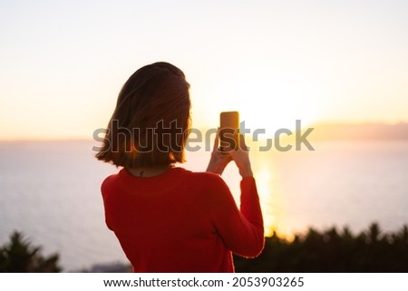 Young woman in a red sweater at a magnificent sunset on the mountain against the background of the sea, photographing the sunset on a mobile phone