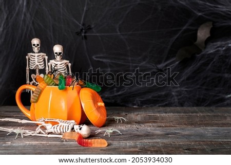 Skeleton toys look into a pumpkin shaped mug filled with festive scary treats, jelly worms and spiders against the cobwebs and dead skeletons. Mystical Halloween dark background, copy space.