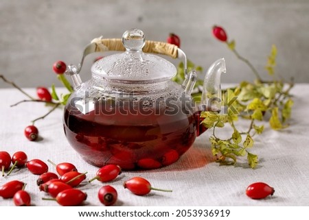 Rose hip berries herbal tea in glass teapot standing on white linen table cloth with wild autumn berries around. Healthy hot drink.