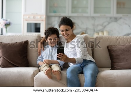 Smiling bonding young Indian woman showing funny mobile applications to adorable little child son, recording streaming online stories for social network, posing for selfie photo, or playing games.
