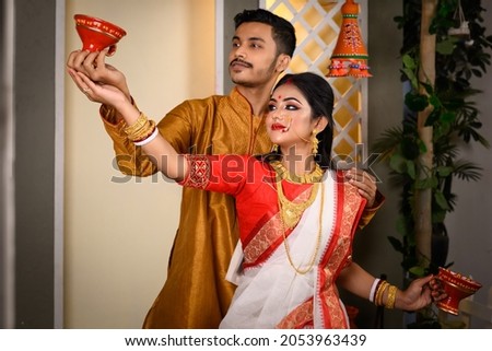 Portrait of Indian man dressed in kurta pyjama with beautiful Indian woman wearing traditional Indian saree, gold jewellery and bangles, holding dhunachi in hands. Royalty-Free Stock Photo #2053963439