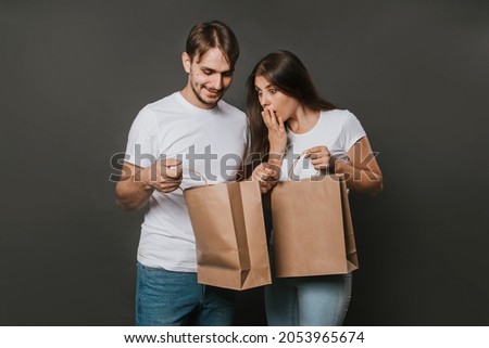 Young man and woman in white blank T-shirts with paper bags in their hands on a solid gray background. Studio photo. Mock-up