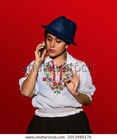 Latin woman in traditional dress and hat, talking through her cell phone with a serious expression in red background