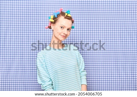 Portrait of happy young blond woman with bright colorful hair curlers on head, with skincare product on face, smiling cheerfully, standing over shower curtain background. Concept of hair and skin care