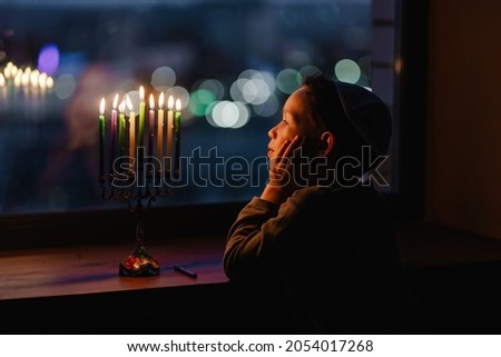 The child lights the menorah for Hanukkah on the windowsill. The boy in the kippah sitting by the window. Jewish holiday. Tradition is a religious ritual. Sunset. The first star. Judaism Royalty-Free Stock Photo #2054017268