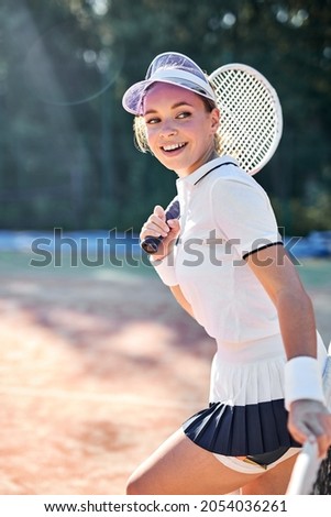 Portrait of young happy cheerful woman in tennis court with racket. Beautiful female tennis player in white uniform and cap stands looking at side, posing. Sports Fashion, People Lifestyle Concept
