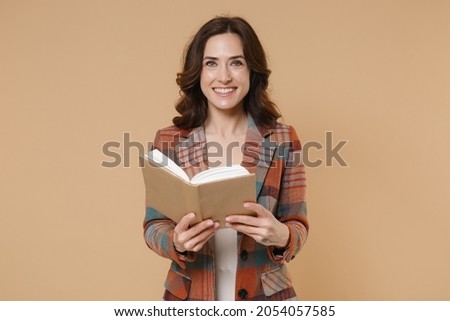 Smiling cheerful attractive young brunette woman 20s wearing casual checkered jacket standing holding in hands reading book looking camera isolated on pastel beige colour background, studio portrait
