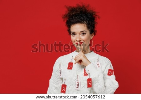 Young smiling happy costumer woman 20s wear white knitted sweater with tags sale in store showroom say hush be quiet with finger on lips shhh gesture isolated on plain red background studio portrait