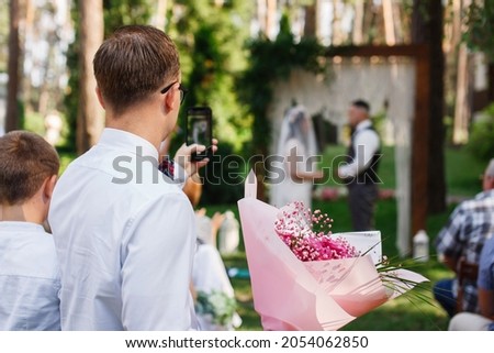 Man  take wedding photo of wedding ceremony on phone. Wedding guest taking photo  bride and groom outdoors in summer. person with phone in hand take photo celebration. Wedding photo on background