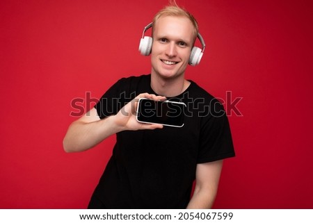 handsome blonde young man wearing black t-shirt and white headphones standing isolated over red background with copy space holding smartphone and showing mobile phone screen listening to music and