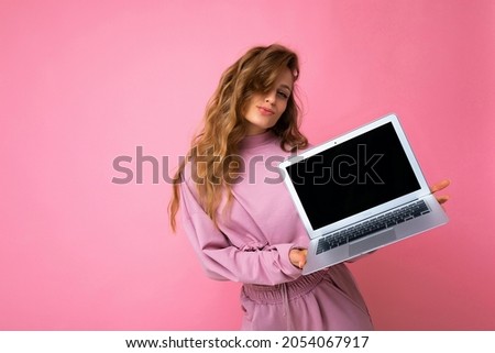 Photo of beautiful blond curly woman holding computer laptop with empty monitor screen with mock up and copy space wearing pink sweater isolated over pink wall background