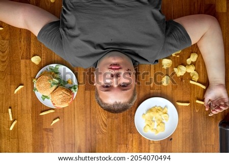 corpulent sad overweight boy lying on floor surrounded by junk food, fries and crisps, sandwich on plate. lazy teenager has no control over eating behavior, tired, after school, leisure time. top view
