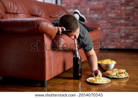 Fat Lazy Boy Taking Fries From Plate Lying On Sofa, Alone At Home, Teenager Boy Having Rest After School, Relaxed. Caucasian child lead unhealthy lifestyle, eating junk meal. overweight, obesity Royalty-Free Stock Photo #2054070545