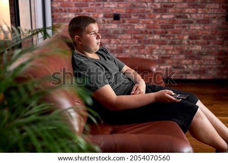 caucasian teenager boy sit on sofa watching tv. Relaxing thoughtful lifestyle portrait in t-shirt looking at television alone after school, lazy and bored. unhealthy lifestyle, need more activity