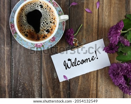 A cup of coffee, flowers and white notepad with text WELCOME wish on a wooden table, flat lay. Space for text
