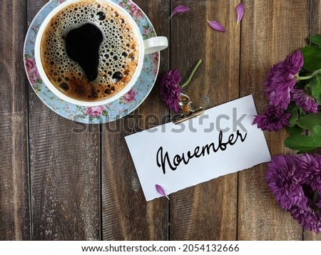 A cup of coffee, flowers and white notepad with text NOVEMBER on wooden table, flat lay. Space for text