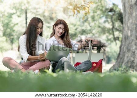 Two young asian university student using computer and talking together with happiness while sitting on green grass under big tree at park.