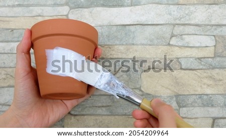Hand holding a new terracotta pot, with another hand holding a white paint brush, making a stroke on the pot. Royalty-Free Stock Photo #2054209964