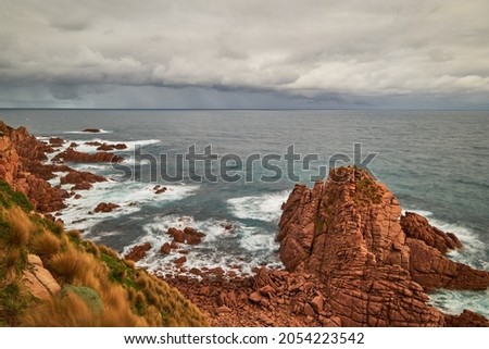 A scenic view overlooking waves breaking against red pillars of rock with the vast ocean on the horizon, in the background is the coastline and sea with cloudy sky above, at Phillip Island Australia.
