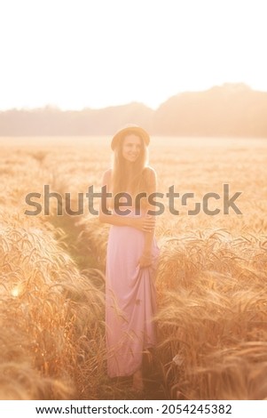 Portrait of a young, happy woman in a straw hat, pink long dress stands in a golden wheat field at sunset. Spikelets of rye in raindrops. Agro walks in the countryside. Copy space textured background