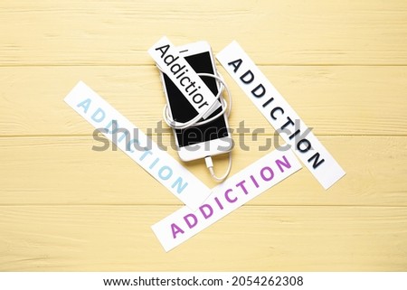 Mobile phone and papers with word ADDICTION on wooden background