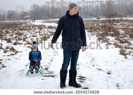 Dad sleds the child in winter, the boy sleds in the forest. Active walks in winter for children. Happy father is pulling child on sled walking on frosty winter day outdoors. 