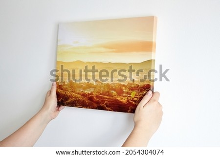 Canvas print with gallery wrap. Woman hangs landscape photography on white wall. Hands holding photo canvas print with image of Barjac in France