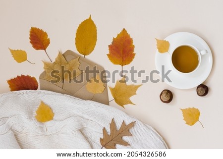 A cup of tea on a white saucer surrounded by fallen autumn leaves and chestnuts, a mail envelope with leaves and a white cozy blanket on a beige background