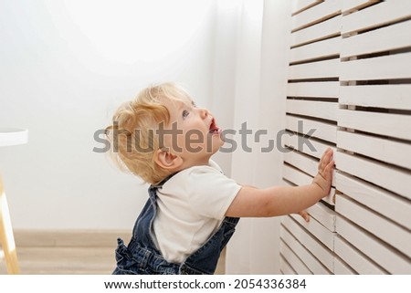 One year old child learning to stand up at living room. Interior background. Adorable blonde little girl in denim overalls leaning on the wall at home. Close up, copy space for text.