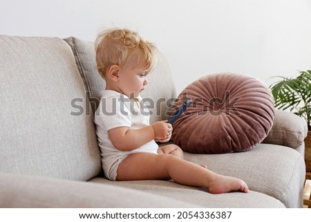 One year old child sitting on the couch, holding the phone and looking at its screen. Adorable blonde little girl watching cartoons on a gadget. Close up, copy space for text, background.