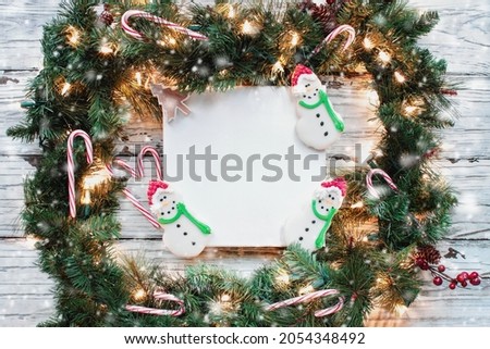 Holiday wreath made of Christmas garland with three Iced Christmas Snowman cookies over a white canvas and rustic wood background. Image shot from flat lay or top view position.