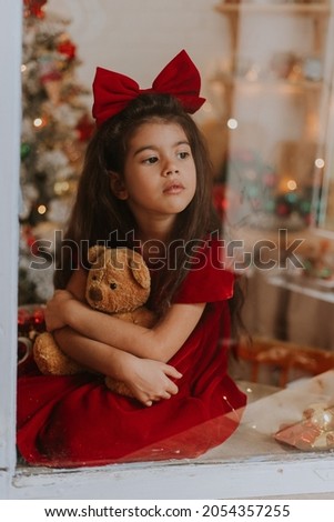 girl in a red dress with a bow on her head with a toy bear is waiting for Santa Claus at the window. close up portrait through glass. space for text. High quality photo