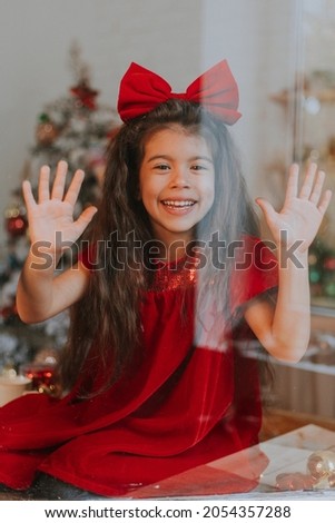 portrait of a little cute girl in a red dress with a bow on her head through glass close up. Merry Christmas and Happy Holidays. Happy New Year. Hygge. Winter. High quality photo
