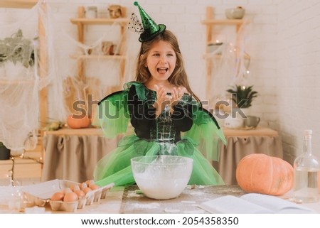 cute little girl in a green Halloween costume of a witch or fairy prepares a pumpkin pie, barm brack in a festive decorated kitchen. space for text. High quality photo