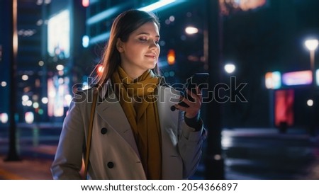 Portrait of a Beautiful Woman in Trench Coat Walking in a Modern City Street with Neon Lights at Night. Attractive Female Using Smartphone and Looking Around the Urban Cinematic Environment. Royalty-Free Stock Photo #2054366897