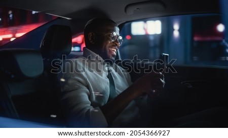 Stylish Black Man in Glasses is Commuting Home in a Backseat of a Taxi at Night. Handsome Male Making a Video Call on a Smartphone while in Transfer Car on City Street with Working Neon Signs.