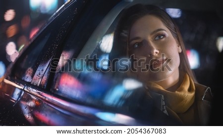 Stylish Female is Commuting Home in a Backseat of a Taxi at Night. Beautiful Woman Passenger Looking Out of Window while in a Car in Urban City Street with Working Neon Signs.